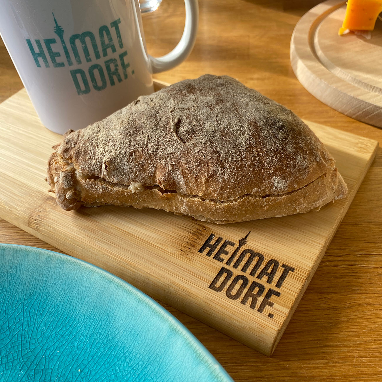 Auf einem hochwertigen Holzbrettchen liegt ein Brötchen und steht eine Tasse mit dem HeimatDorf Logo Düsseldorf. Das Frühstücksbrettchen hat unten rechts eine Lasergravur mit dem Heimatdorf Logo. Das i ist durch den Rheinturm dargestellt.